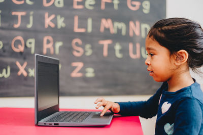 Side view of diverse pre school age girl at home doing distance learning on laptop computer
