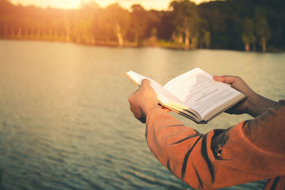 Midsection of man holding book against sea during sunset