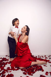 Mother in a red dress is sitting on the floor with a child,  son in rose petals on valentine's day