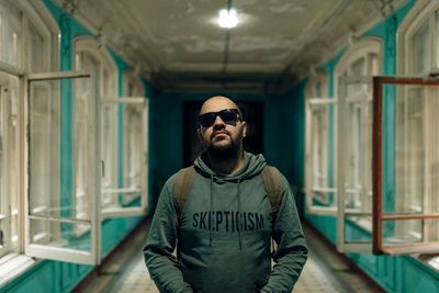 Portrait of man wearing sunglasses standing amidst open windows in corridor