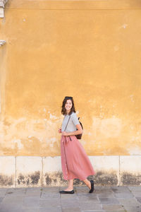 Smiling young woman walking on footpath against wall