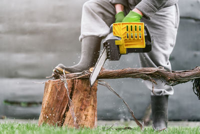 Close up of man cutting a log with a chainsaw in the garden leaning on another log on a sunny day