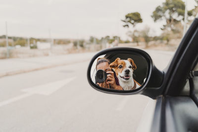 Reflection of woman photographing by dog in side-view mirror car