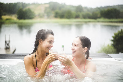 Smiling female friends toasting wineglasses while enjoying in hot tub against lake