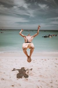 Full length of shirtless man jumping at beach against sky