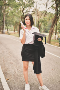 Young woman holding mortarboard and graduation gown on street