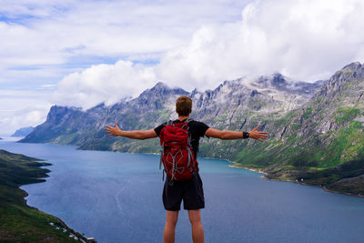 Rear view of man with arms outstretched standing at lakeshore against mountains