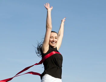 Low angle portrait of cheerful woman with arms raised against blue sky