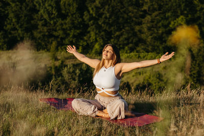 Young woman sunbathing and meditating on the mat outdoors at sunset with beautiful landscape
