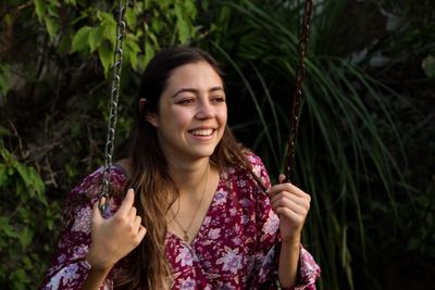 Smiling young woman looking away sitting on swing against plants
