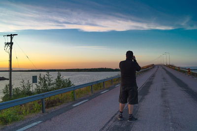 Rear view of man photographing standing on road during sunset