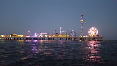 Illuminated amusement park by sea against clear sky at night
