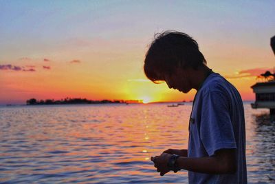 Side view of mature man using mobile phone while standing by sea against sky during sunset