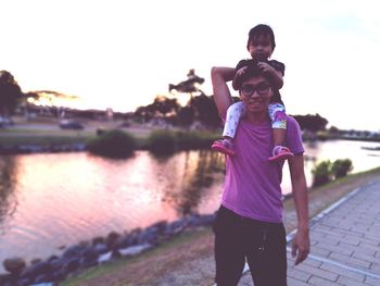 Portrait of smiling father carrying daughter on shoulders by river during sunset