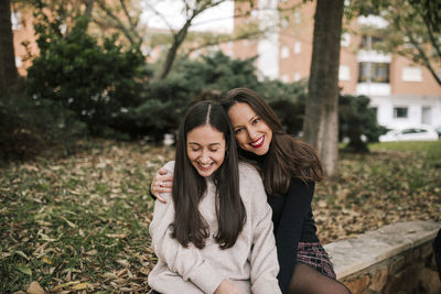 Smiling mother with daughter sitting on retaining wall in park