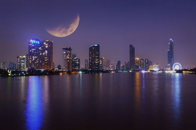 Sea by illuminated modern buildings against sky at night