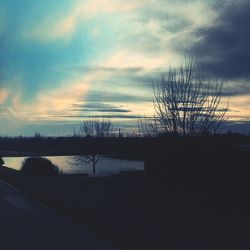 Bare trees on landscape against cloudy sky