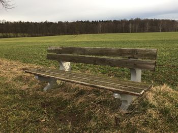 Empty bench on field by trees against sky