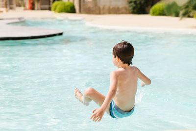 Back view of unrecognizable shirtless little boy in shorts jumping into outdoor swimming pool during summer holidays and having fun