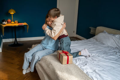 Happy little son hugging mother on bed at home, going to unpack festive wrap. birthday concept.