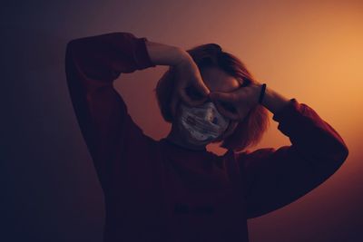 Portrait of young woman wearing mask gesturing while standing against brown background