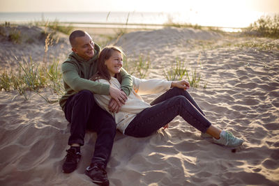 Husband and wife in hoodies and sneakers sitting on a sandy beach by the sea in autumn