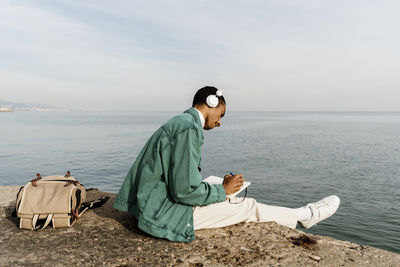 Young man with headphones writing in diary by sea and sky