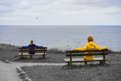 Rear view of people sitting on bench by sea against sky