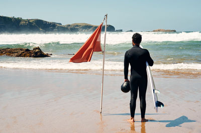 Back view full length of anonymous man wearing surfing suit standing near waving flag on coast washed by foamy waves