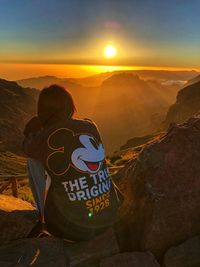 Rear view of man sitting on rock against sky during sunset