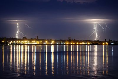 Lightning over illuminated city by sea against sky at night