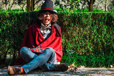 Man cosplaying mad hatter sitting on ground against hedges