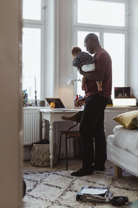 Businessman carrying baby boy while using laptop at home office