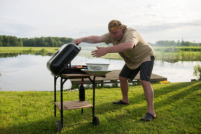 Young man grilling by lake against sky