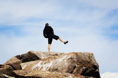 Rear view of man standing on one leg at rock against sky