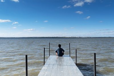 Rear view of man sitting on pier by river against blue sky during sunny day