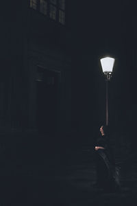 Man sitting in illuminated street light against building at night