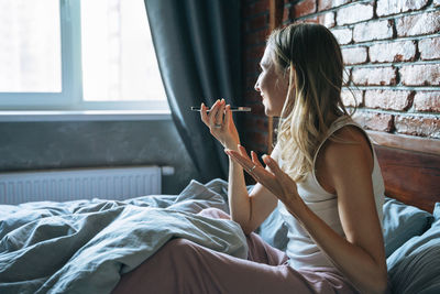 Adult woman with blonde long hair in pajamas using mobile phone on bed in loft room at home