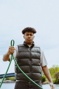 Low angle portrait of confident young man holding garden hose against sky