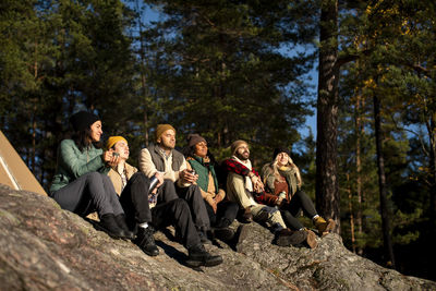 Multiracial male and female friends enjoying sunlight while sitting on rock in forest