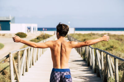 Rear view of shirtless boy standing by railing against sky