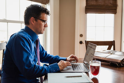 Man in shirt and tie working from home using computer at dining table.