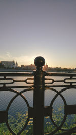 Metal railing by river against sky during sunset