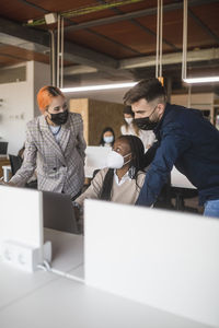 Group of diverse coworkers in protective masks gathering on laptop and discussing project while working together in contemporary workspace