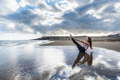 Young woman exercising at beach against cloudy sky