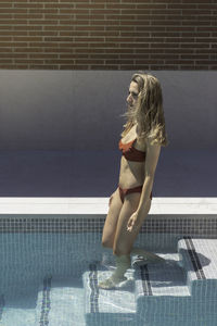 Side view of young blonde woman on bikini entering the water on the swimming pool in her house