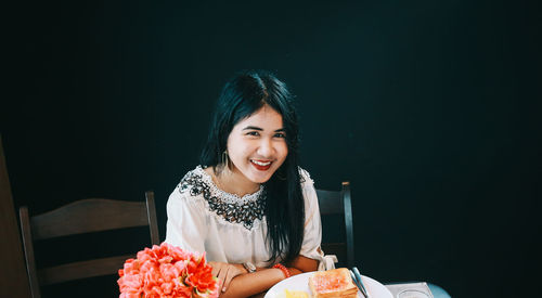 Portrait of smiling woman with breakfast on table sitting against black background