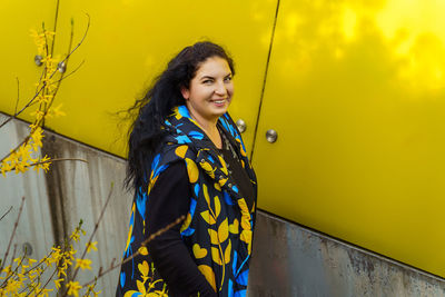 Happy ukrainian woman in blue and yellow waistcoat walking along yellow wall and blooming forsythia