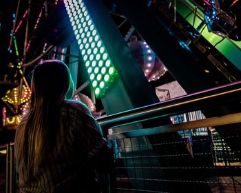Rear view of woman standing by illuminated lighting equipment and railing at night