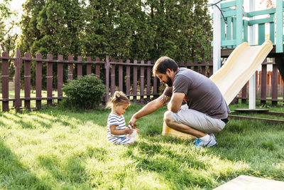 Father playing with daughter sitting on grassy field at backyard